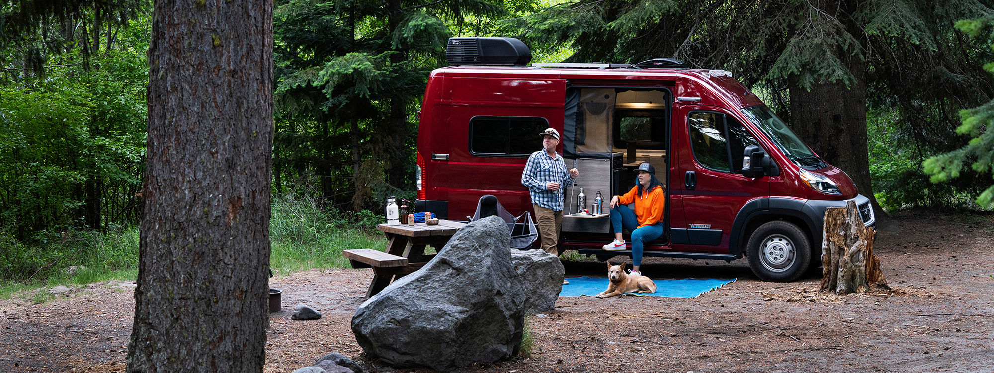 Young couple sits outside the Winnebago Solis Pocket enjoying the surrounding forest